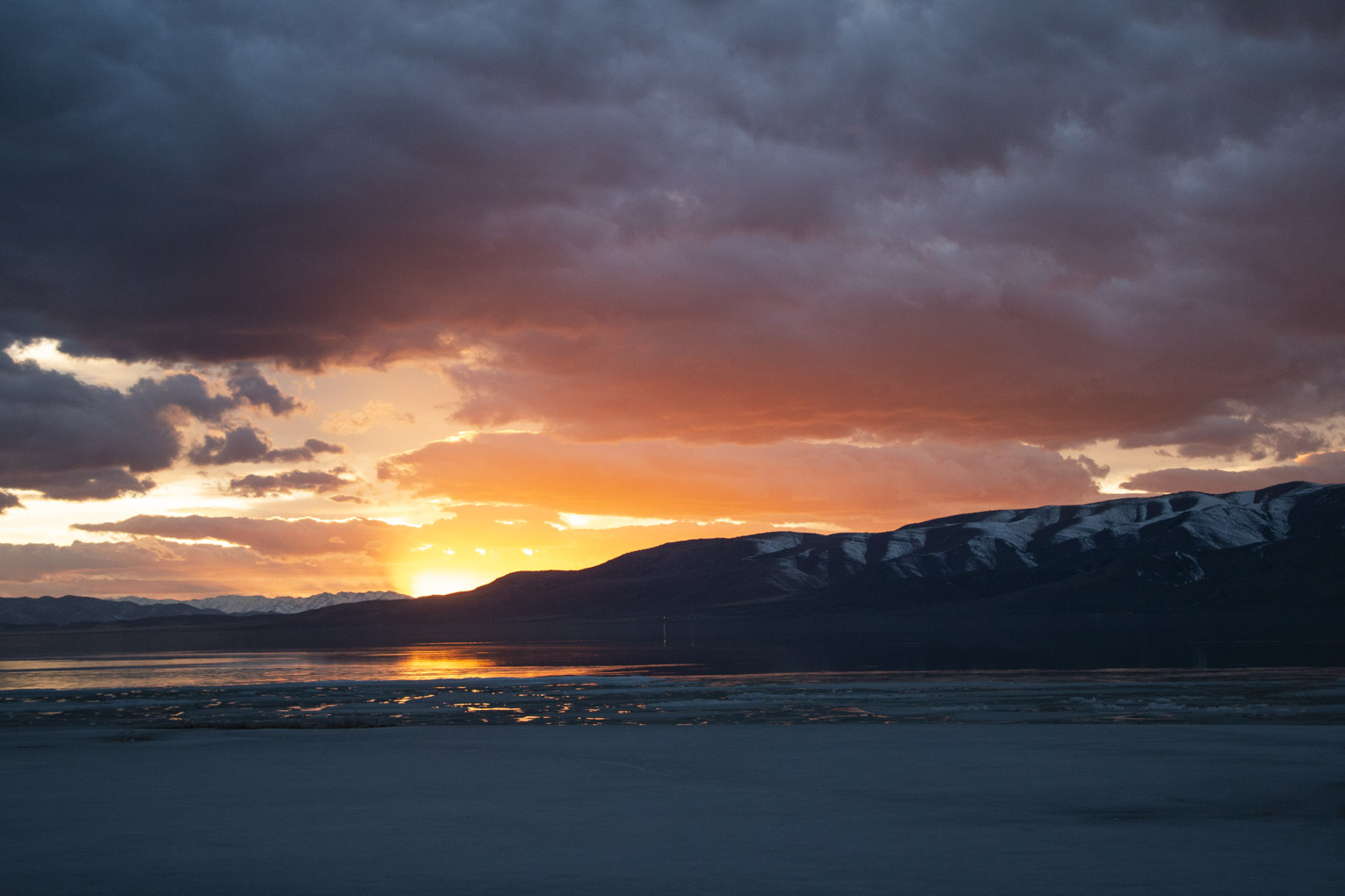 The sun sets just behind dark blue mountains throwing a flare of light up reflected in the lake until it turns to ice